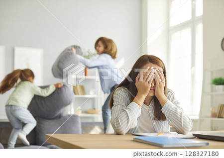 Exhausted mother sitting at desk with head in hands as kids jumping and playing loudly on sofa. 128327180