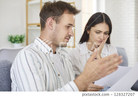 Portrait of young frustrated couple sitting on sofa looking through financial documents at home 128327207