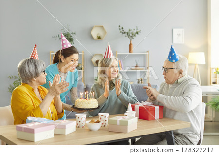Happy seniors celebrating birthday with cake and presents, enjoying festive moment with young nurse. 128327212