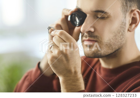 Young man examining diamond ring through magnifying loupe, analyzing gemstone quality and clarity. 128327233