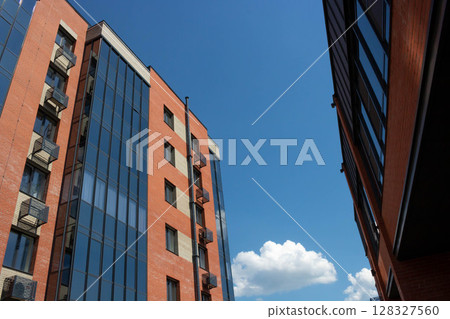 Against the backdrop of a blue sky, a red brick building with a glass facade reflects a blue sky with white clouds. Against the backdrop of a blue sky, a red brick building with a glass facade reflects a blue sky with white clouds. 128327560
