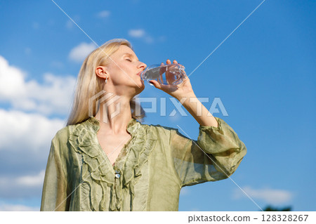 Refreshing Hydration: A Woman Enjoys a Bottle of Water Under a Bright Blue Sky with Fluffy Clouds, Highlighting the Importance of Staying Hydrated during Warm Days and Outdoor Activities 128328267