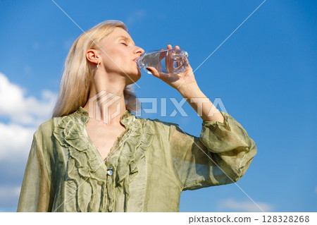 Refreshing Hydration: A Woman Enjoying Water Under a Bright Blue Sky in a Soft Green Blouse with Light Ruffled Details in Nature's Embrace 128328268