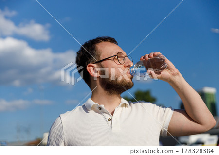 A Young Man Enjoying Refreshing Water Outdoors on a Bright Day with a Clear Blue Sky, Emphasizing Health and Hydration in a Casual Setting 128328384