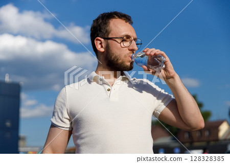 Man Quenching Thirst: Stylish Individual Enjoying Refreshing Bottled Water Under Clear Blue Sky on a Bright Day with Cloudy Background in Urban Setting Man Quenching Thirst: Stylish Individual Enjoying Refreshing Bottled Water Under Clear Blue Sky on a Bright Day with Cloudy Background in Urban Setting 128328385