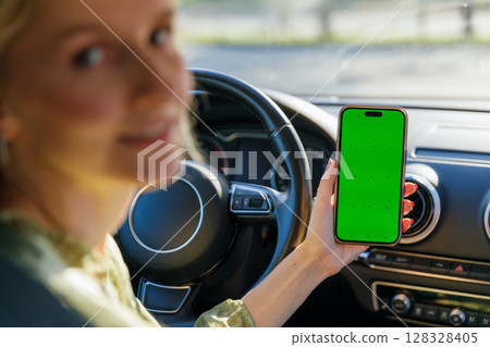 Smiling Woman in Car Displaying Smartphone with Green Screen While Driving, Perfect for Template Use in Advertisements and Digital Content Creation 128328405