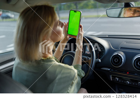 Woman in a Car Holding a Smartphone with Green Screen While Driving: A Glimpse Into Multi-tasking and Technology's Role in Modern Commuting 128328414