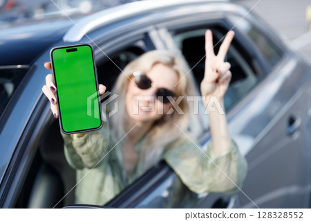 Cheerful Woman Showing Green Phone Screen While Leaning Out of Car Window with Peace Sign Gesture in a Bright Outdoor Setting Cheerful Woman Showing Green Phone Screen While Leaning Out of Car Window with Peace Sign Gesture in a Bright Outdoor Setting 128328552