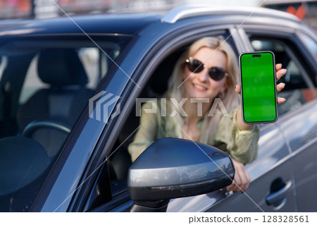 A Young Woman with Long Hair and Sunglasses Holds Up a Smartphone with a Green Screen from a Car Window, Radiating Joy and Modern Connectivity, Perfect for Display and Sharing 128328561