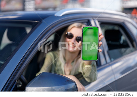 Cheerful Woman Displays Smartphone with Green Screen from Car Window, Showcasing Technology and Connection While Enjoying a Beautiful Day Outdoors 128328562