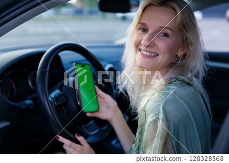 A Young Woman in a Car Holding a Smartphone with a Green Screen, Smiling Cheerfully, Capturing the Joy of Modern Technology and Life 128328568