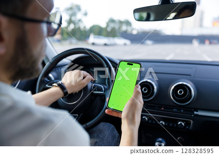 A man inside a car using a smartphone with a vibrant green screen, showcasing the intersection of technology and driving in a modern urban setting with clear concentration. A man inside a car using a smartphone with a vibrant green screen, showcasing the intersection of technology and driving in a modern urban setting with clear concentration. 128328595