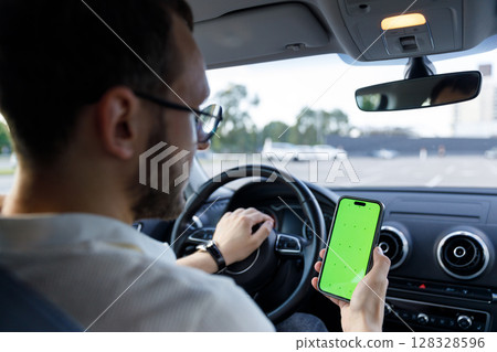 Man in a Car Holding a Smartphone with a Green Screen While Driving Thoughtfully Contemplating Ahead in a Parked Environment 128328596