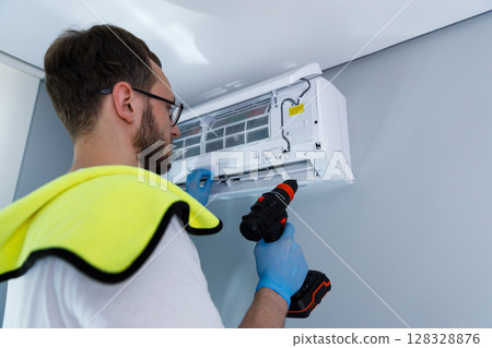 Professional technician performs routine maintenance on a modern air conditioning unit mounted on a wall, ensuring optimal performance in a comfortable indoor environment. Professional technician performs routine maintenance on a modern air conditioning unit mounted on a wall, ensuring optimal performance in a comfortable indoor environment. 128328876