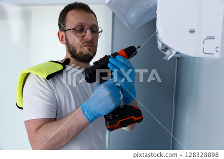 Man in Blue Gloves Holding a Power Drill While Preparing for Installation Tasks, Wearing a White T-Shirt with a Bright Yellow Towel on His Shoulder Indoors in a Modern Setting Man in Blue Gloves Holding a Power Drill While Preparing for Installation Tasks, Wearing a White T-Shirt with a Bright Yellow Towel on His Shoulder Indoors in a Modern Setting 128328898