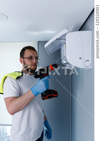 Man Utilizing a Power Drill for Installing a Wall-Mounted Air Conditioning Unit in a Modern Indoor Space with Safety Gloves and a Towel on His Shoulder Man Utilizing a Power Drill for Installing a Wall-Mounted Air Conditioning Unit in a Modern Indoor Space with Safety Gloves and a Towel on His Shoulder 128328901