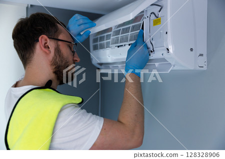 A dedicated technician performing maintenance on an air conditioning unit, carefully removing components while ensuring cleanliness and safety with blue gloves and yellow vest, showcasing attention to A dedicated technician performing maintenance on an air conditioning unit, carefully removing components while ensuring cleanliness and safety with blue gloves and yellow vest, showcasing attention to 128328906