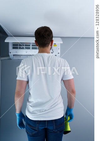 A Person Wearing Blue Gloves and a White T-shirt Preparing to Clean an Air Conditioner Unit in a Gray Room with Modern Design Elements and Minimalist Aesthetic Features 128328936