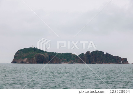 A Majestic Coastal Landscape Featuring a Rocky Island and a Lighthouse Amidst a Cloudy Sky with Muted Colors and Calm Waters, Perfect for Seascape Photography Enthusiasts A Majestic Coastal Landscape Featuring a Rocky Island and a Lighthouse Amidst a Cloudy Sky with Muted Colors and Calm Waters, Perfect for Seascape Photography Enthusiasts 128328994