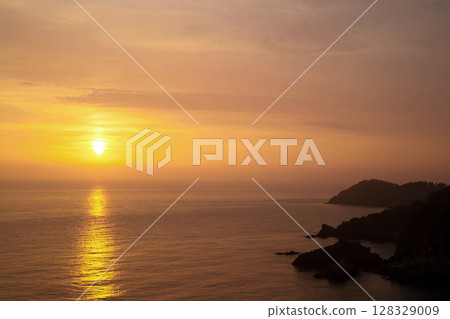 Spectacular Sunset Over Calm Waters: A Breathtaking View of the Sun Setting on the Horizon at Dusk Behind Serene Ocean Waves and Rocky Shorelines 128329009