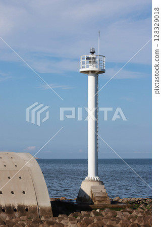 Coastal Beacon Standing Tall Over the Serene Waters with White Cloudy Sky and Rocky Jetty in the Foreground Reflecting a Beautiful Day by the Sea 128329018