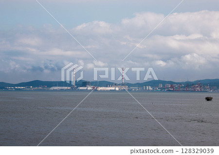 A panoramic view of a coastal industrial landscape featuring a prominent power plant, smokestacks, and heavy machinery along the serene waters against the backdrop of distant mountains. A panoramic view of a coastal industrial landscape featuring a prominent power plant, smokestacks, and heavy machinery along the serene waters against the backdrop of distant mountains. 128329039