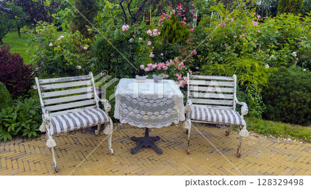 Charming Garden Setting with Rustic White Benches and a Lace-Covered Table Surrounded by Lush Roses and Greenery Creating a Serene Outdoor Retreat for Relaxation 128329498