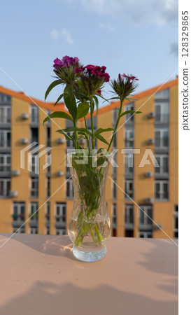 Stunning Floral Display in a Transparent Vase on a Sunny Balcony with Apartment Building in the Background, Capturing a Serene Urban Vibe Amidst Nature's Beauty Stunning Floral Display in a Transparent Vase on a Sunny Balcony with Apartment Building in the Background, Capturing a Serene Urban Vibe Amidst Nature's Beauty 128329865