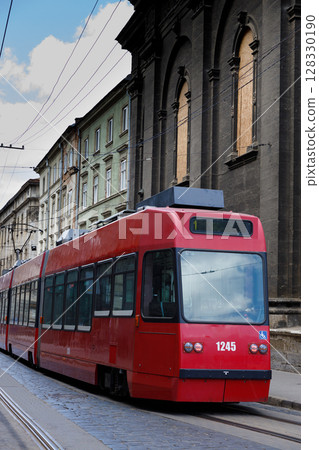 A bright red tram making its way through a historic city street, featuring old buildings and clear blue skies, showcasing the blend of modern transportation and traditional architecture. 128330190