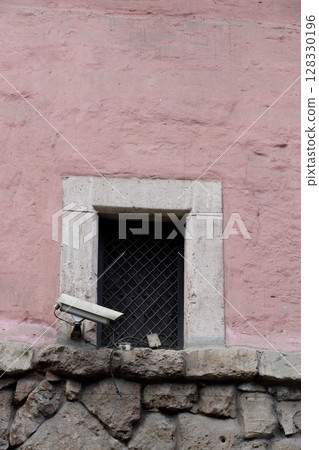 Surveillance Camera Overlooking a Shaded Window Set Against a Pink Wall with Stone Base: A Blend of Modern Security and Historical Architecture's Repose Surveillance Camera Overlooking a Shaded Window Set Against a Pink Wall with Stone Base: A Blend of Modern Security and Historical Architecture's Repose 128330196