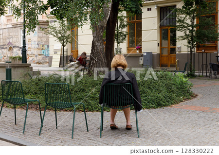 A Serene Outdoor Scene Featuring a Woman Sitting Back on a Chair in a Quaint European Street Surrounded by Lush Greenery and Architectural Beauty A Serene Outdoor Scene Featuring a Woman Sitting Back on a Chair in a Quaint European Street Surrounded by Lush Greenery and Architectural Beauty 128330222