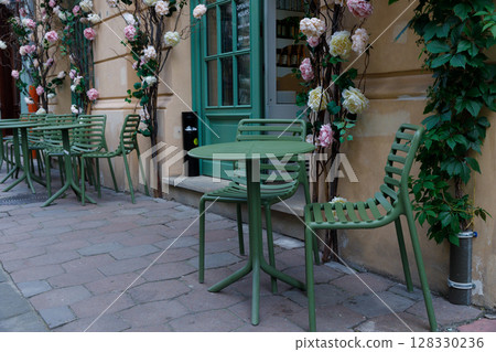 Charming Outdoor Cafe Setting with Green Chairs and Floral Decorations by the Entrance, Perfect for Relaxing Moments and Coffee Breaks in a Picturesque Atmosphere Charming Outdoor Cafe Setting with Green Chairs and Floral Decorations by the Entrance, Perfect for Relaxing Moments and Coffee Breaks in a Picturesque Atmosphere 128330236