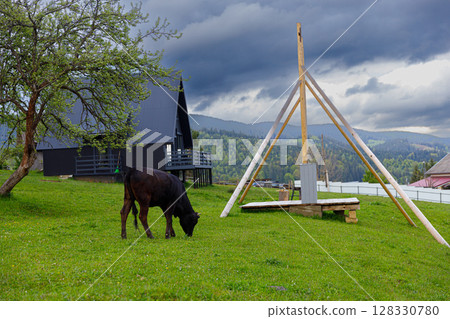 A Peaceful Rural Scene Featuring a Grazing Cow, Green Pasture, and Simple Structures Under Dramatic Cloudy Skies - An Idyllic Retreat in Nature's Embrace A Peaceful Rural Scene Featuring a Grazing Cow, Green Pasture, and Simple Structures Under Dramatic Cloudy Skies - An Idyllic Retreat in Nature's Embrace 128330780
