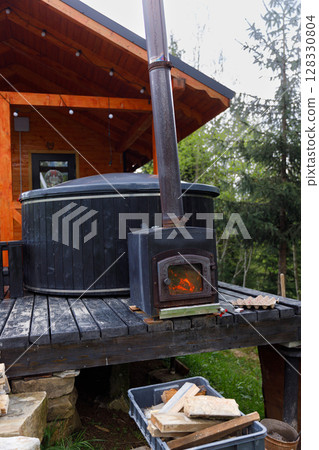 Rustic Outdoor Setup Featuring Wood-Burning Stove Alongside Hot Tub on Wooden Deck Surrounded by Lush Greenery Under Partially Cloudy Sky 128330804