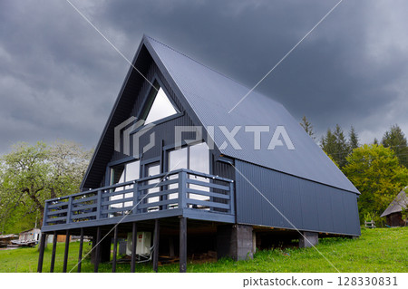 Modern A-Frame House Surrounded by Greenery Under a Dramatic Cloudy Sky, Showcasing Contemporary Design with Large Windows and Elevated Deck for Outdoor Enjoyment 128330831