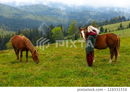 A Serene Moment in Nature: A Young Individual Interacting with Two Horses Surrounded by Lush Green Mountains and Scenic Wilderness Landscape A Serene Moment in Nature: A Young Individual Interacting with Two Horses Surrounded by Lush Green Mountains and Scenic Wilderness Landscape 128331558