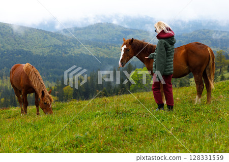 A Serene Moment: A Girl Interacting with Horses in a Picturesque Mountain Landscape Surrounded by Lush Grass and Scenic Beauty, Perfect for Nature Lovers and Equestrian Enthusiasts A Serene Moment: A Girl Interacting with Horses in a Picturesque Mountain Landscape Surrounded by Lush Grass and Scenic Beauty, Perfect for Nature Lovers and Equestrian Enthusiasts 128331559