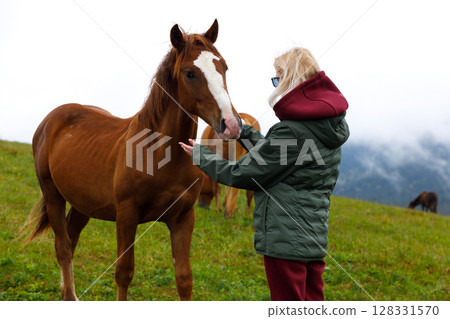 A Young Girl Interacting with a Beautiful Brown Horse in a Lush Green Field Under a Cloudy Sky, Representing the Bond Between Humans and Nature in Tranquil Surroundings A Young Girl Interacting with a Beautiful Brown Horse in a Lush Green Field Under a Cloudy Sky, Representing the Bond Between Humans and Nature in Tranquil Surroundings 128331570