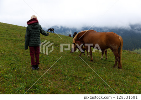 A Young Child Interacting with Horses on a Scenic Hillside Amidst Cloudy Mountains and Lush Green Fields, An Ideal Representation of Nature and Childhood Innocence 128331591