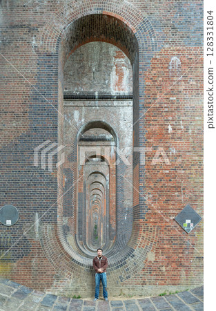 View of a young man wearing a black T-shirt and jeans standing on the structure under the bridge of Ouse Valley Viaduct or Balcombe Viaduct. 128331804