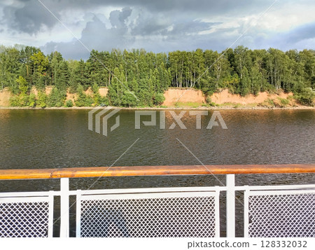 Dramatic scene of dark thunderclouds over a pine forest on a riverbank, viewed from the side of a ship 128332032