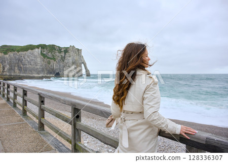 Woman in beige trench looking at Etretat cliff in Normandy Woman in beige trench looking at Etretat cliff in Normandy 128336307