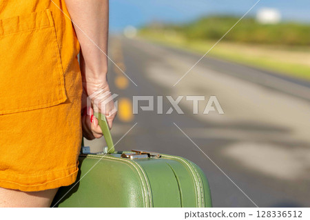 Woman's hand is holding a retro green suitcase and walking on a highway. 128336512