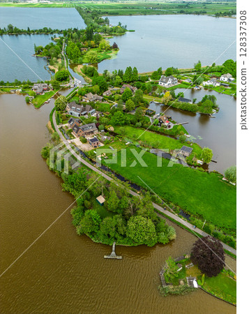 Aerial view of a scenic lakeside village with narrow peninsulas, green fields, and houses surrounded by calm waters under a cloudy sky in the Dutch countryside. Aerial view of a scenic lakeside village with narrow peninsulas, green fields, and houses surrounded by calm waters under a cloudy sky in the Dutch countryside. 128337308