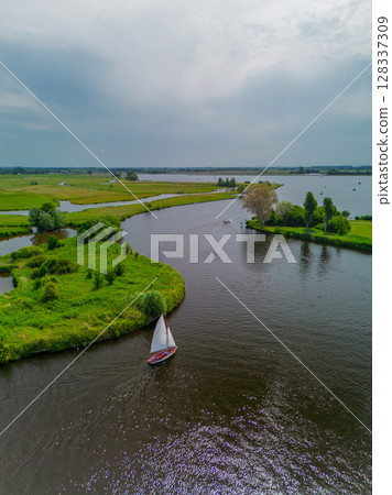Aerial view of a sailboat cruising through a winding river surrounded by lush green wetlands and trees under a moody, cloud-covered sky in a peaceful countryside landscape. Aerial view of a sailboat cruising through a winding river surrounded by lush green wetlands and trees under a moody, cloud-covered sky in a peaceful countryside landscape. 128337309