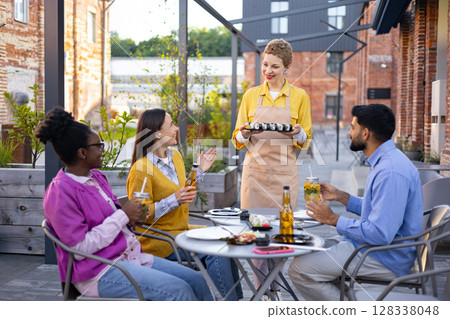 A group of friends enjoying sushi and drinks at an outdoor restaurant, served by a friendly waitress. 128338048