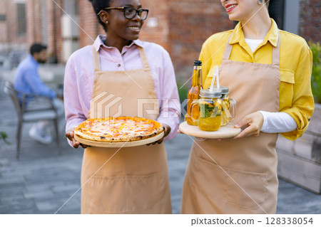 Two waitresses smiling and holding food and drinks outdoors at a restaurant. 128338054