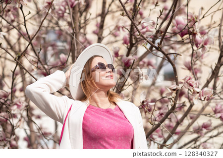 Magnolia Spring Woman: Enjoys blossoms outdoors daytime in city, finds beauty, wears hat, sunglasses. Magnolia Spring Woman: Enjoys blossoms outdoors daytime in city, finds beauty, wears hat, sunglasses. 128340327