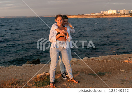 Couple Beach Sunset Hug - Two people hug while overlooking the ocean at sunset. They are on a rocky beach. The picture was likely taken during a vacation or romantic getaway. 128340352