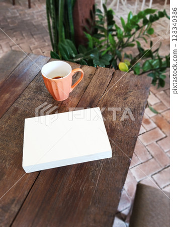 An unidentified book with a white cover placed next to a beige ceramic teacup on a wooden table. 128340386